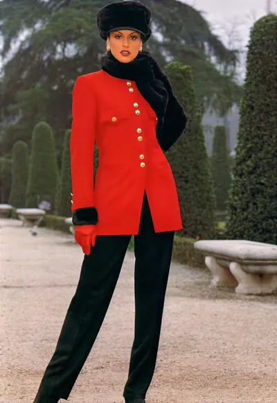 This appears to be a fashion editorial or advertisement photograph from the late 1980s or early 1990s, shot outdoors in a formal garden with clipped topiary hedges and stone benches. A woman with dark hair poses wearing a bright red military-style jacket with gold buttons down the front and black fur trim at the cuffs, black fitted trousers, a black fur stole draped over one shoulder, a black fur pillbox-style hat, and red leather gloves.