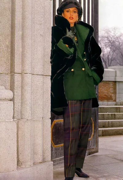 A fashion magazine or advertisement photograph from what appears to be the 1980s, showing a woman with dark hair wearing a dark fur-trimmed black coat draped over her shoulders, a forest green double-breasted wool blazer with gold buttons, dark plaid trousers in navy, green, red, and yellow, dark green leather gloves, a dark fur-trimmed beret-style hat, and gold earrings. She is posed beside a stone column near ornate iron gates, with stone steps and bare trees visible in the background.
