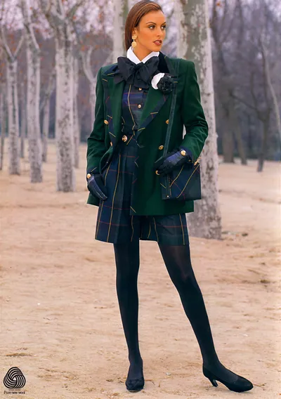 This is a fashion advertisement, marked with a Pure New Wool logo in the lower left corner. A woman with brown hair pulled back is standing outdoors among bare trees on sandy ground, wearing a double-breasted dark green blazer with gold buttons, a tartan plaid skirt and matching vest or dress underneath in navy, green, and red plaid, a white collared shirt with a large black bow at the neck, a black fabric flower brooch, black leather gloves, black opaque tights, and black low-heeled pumps.