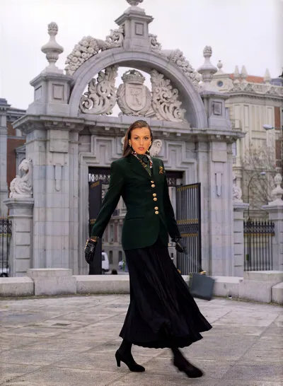 This appears to be a fashion magazine editorial or advertisement photograph from the late 1980s or early 1990s, showing a woman with brown hair walking in front of an ornate stone archway gate with heraldic carvings, likely in Madrid, Spain based on the architecture. She is wearing a dark forest green blazer jacket with gold buttons, a patterned silk scarf at the neck, gold earrings, black leather gloves with decorative cuffs, a black pleated midi skirt, black hosiery, and black heeled pumps.
