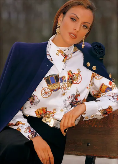 This appears to be a fashion advertisement or magazine spread featuring a woman with brown hair wearing a white satin blouse printed with colorful horse-drawn carriages and equestrian motifs in gold, red, and blue, paired with a separate solid navy blue blazer draped over her shoulders that has gold buttons and a fabric rosette at the shoulder. She also wears gold drop earrings, a wide metallic gold belt, black trousers, and is posed leaning against a wooden surface outdoors.