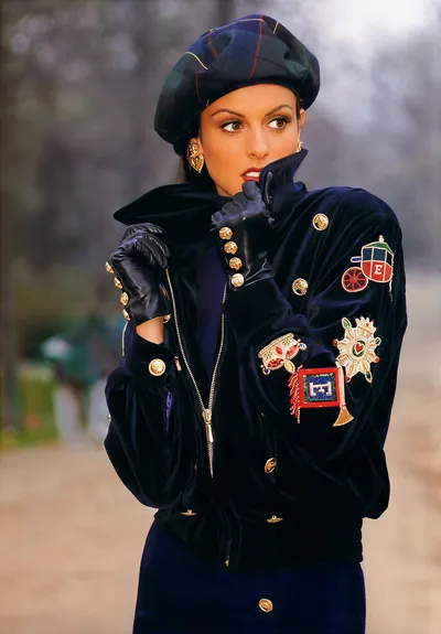 A fashion magazine or advertisement photograph of a woman with brown hair and brown eyes, wearing a navy blue velvet zip-up bomber jacket heavily embellished with gold coin buttons and colorful beaded appliqués depicting a carriage, a flag, and a starburst medallion. She wears matching navy blue leather gloves with gold buttons, gold heart-shaped earrings, and a dark green and navy blue tartan plaid beret. The background is an outdoor setting with blurred trees.