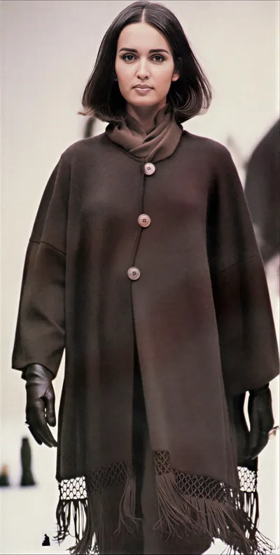 A runway photo of a woman with short dark hair wearing an oversized dark brown coat with three large buttons, a draped cowl neck, fringed hem, and black leather gloves.
