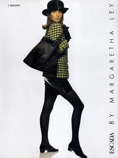 This is a vintage fashion advertisement for Escada by Margaretha Ley, sold at I. Magnin. A woman with shoulder-length brown hair poses in profile against a plain white background, looking back over her shoulder. She wears a black bowler hat, a yellow and black houndstooth-patterned oversized jacket, a black patent leather mini skirt, black opaque tights, and black low-heeled flat shoes. She carries a large black quilted leather tote bag over one shoulder and holds black gloves in her hand.