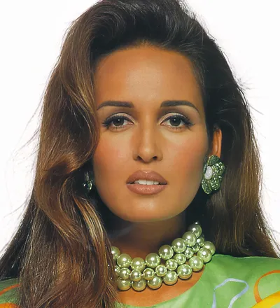 A close-up portrait photograph of a woman with long brown hair, wearing green floral clip-on earrings, a multi-strand necklace of large pale green pearl-like beads, and a green patterned top or scarf. The background is plain white.