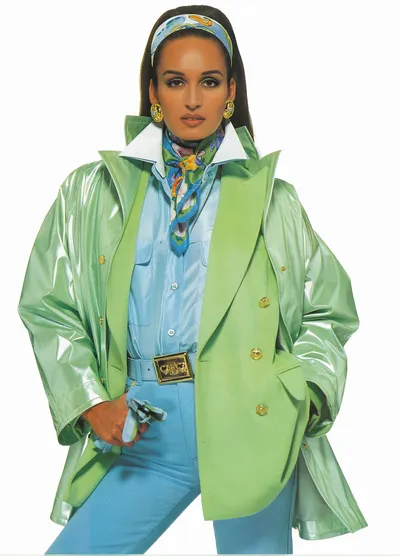 A fashion advertisement or magazine editorial photograph featuring a woman with dark hair pulled back, wearing a colorful layered outfit. She is dressed in a shiny metallic light green oversized raincoat or vinyl jacket worn open over a matte light green double-breasted blazer with gold buttons, a light blue satin button-up shirt, light blue trousers with a gold decorative belt buckle, and a multicolored floral silk scarf tied loosely around her neck. She also wears a matching patterned headband, gold earrings, and holds light blue gloves in one hand.
