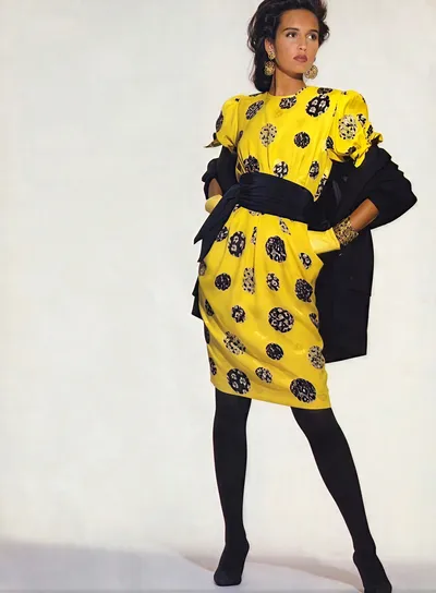 A fashion editorial or magazine spread photograph showing a woman with dark hair wearing a bright yellow short-sleeve dress printed with black-and-navy circular floral medallions, cinched at the waist with a wide dark sash, paired with black tights and a black blazer draped over her shoulders, along with gold hoop earrings and a gold cuff bracelet.