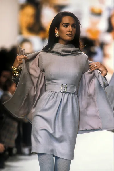 This is a runway photo of a woman with long dark hair wearing a pale grey-blue belted mini dress with a cowl neckline and ribbed light blue tights, while holding open a matching grey-blue oversized coat with wide sleeves. She is also wearing gold hoop earrings and a gold cuff bracelet, and is walking on a white runway with a blurred audience visible in the background.