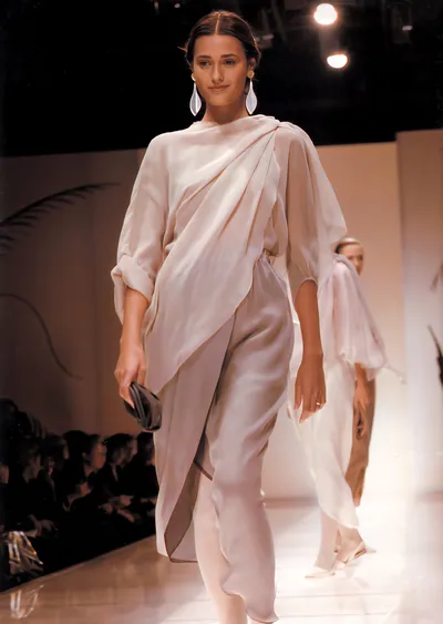 A runway photo showing a woman with dark hair pulled back, wearing a draped floor-length ivory/cream chiffon dress with three-quarter sleeves and long white drop earrings, holding a small dark clutch; a second model in a similar pale outfit is visible behind her on the lit catwalk, with a seated audience in the background.