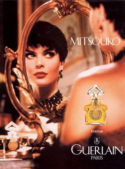 This is a vintage perfume advertisement for Mitsouko by Guerlain Paris. It shows a woman with short dark hair, wearing dark lipstick and a black beaded neckpiece, gazing into an ornate gold-framed mirror, with the decorative amber glass perfume bottle displayed in the foreground.