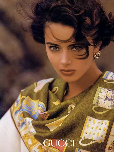 A Gucci advertisement featuring a close-up portrait of a young woman with short, dark curly hair, wearing gold chain-link clip earrings and a large olive green silk scarf with a decorative printed pattern in gold, blue, white, and yellow draped over a white top.
