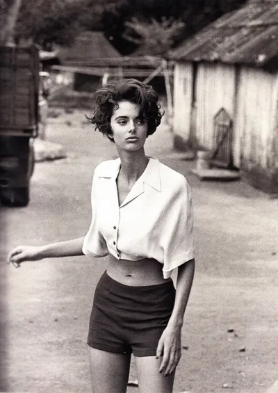 A black-and-white fashion photograph showing a young woman with short curly dark hair, wearing an open collared short-sleeve shirt tied above the waist and high-waisted shorts, standing on a dirt road with rustic wooden buildings in the background.