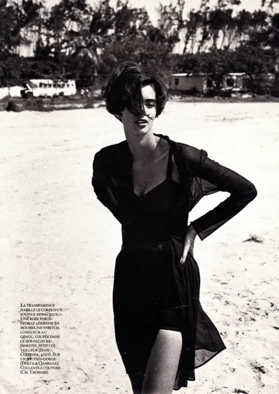 A black-and-white fashion magazine spread showing a woman with short dark hair wearing a sheer black wrap dress, standing on a sandy beach with trees and buildings visible in the background. French caption text in the lower left describes the clothing items featured.
