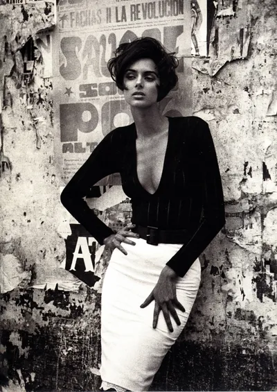 A black-and-white fashion photograph of a woman with short dark hair wearing a deep V-neck long-sleeve black top, a wide black belt, and a fitted white pencil skirt, posing against a weathered wall covered in torn Spanish-language posters.