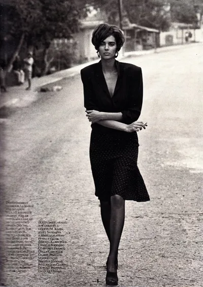 A black-and-white magazine editorial photo showing a woman with short dark hair wearing a dark blazer and a dotted midi skirt, walking on an outdoor street or path. French fashion credits are printed along the left and bottom edges of the image.