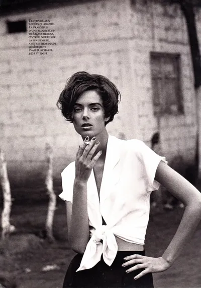 A black-and-white magazine fashion spread showing a woman with short dark hair wearing a white tied blouse and dark skirt, holding a cigarette, standing in front of a weathered stone wall. French text in the upper left describes the Cacharel clothing items featured.