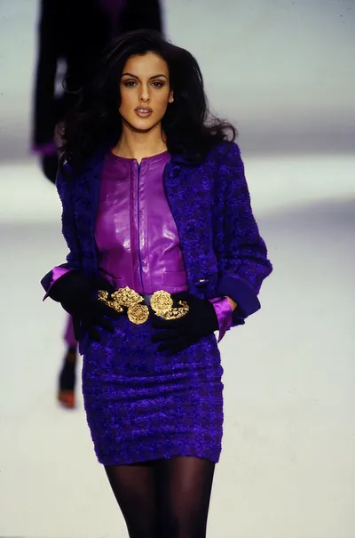 This is a runway photo of a woman with long dark wavy hair walking on a white catwalk. She wears a deep royal blue textured boucle-style jacket over a magenta or bright purple leather zip-front top, paired with a matching royal blue textured mini skirt, a wide black belt with an ornate gold buckle, black gloves with purple satin cuffs, and dark sheer tights.