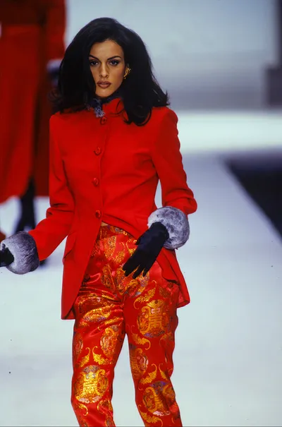 This is a runway photo of a woman with dark shoulder-length hair wearing a structured red jacket with a fur-trimmed collar and black gloves with gray fur cuffs, paired with red satin pants printed with an ornate gold and multicolor Asian-inspired pattern. She is walking on a white runway.
