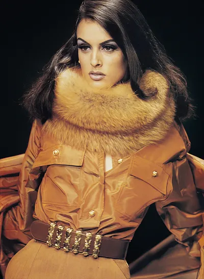 A fashion runway or editorial photograph of a woman with long dark hair wearing a copper/amber-toned satin trench coat with gold buttons, a large fur collar, and a wide brown belt with a gold buckle, posed against a dark background.
