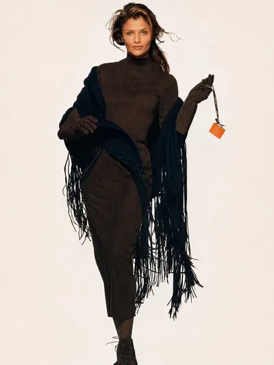 This is a fashion editorial or advertisement photograph featuring a woman with brown hair wearing a dark brown ribbed turtleneck midi dress and dark brown long gloves. She is draped in a large navy blue fringed shawl or wrap, and she holds a small orange cube-shaped accessory or bag charm dangling from her raised gloved hand. She is also wearing dark ankle boots.