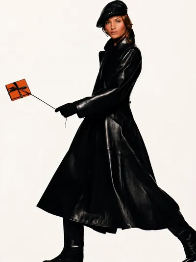This is a fashion advertisement featuring a woman with brown hair wearing a long black leather coat, black leather gloves, black knee-high boots, and a black leather beret-style cap. She is mid-stride against a plain white background, holding a small orange gift box tied with a black ribbon by a thin string.