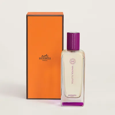 This is a product advertisement photograph showing a bottle of Hermes Paris perfume called Violette Volynka from the Collection Hermessence, with a clear glass bottle, a purple leather cap with white stitching, and a purple base, displayed in front of the brand's signature orange box with brown trim.