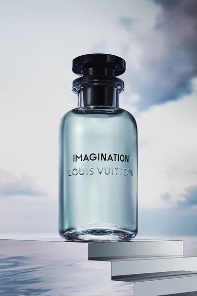 This is a product advertisement photograph showing a glass perfume bottle with a light blue-green tint and a black stopper cap, embossed with the words IMAGINATION and LOUIS VUITTON, displayed on stacked metallic rectangular platforms against a sky and water background.