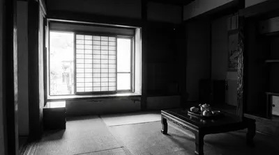 A black-and-white interior photograph of a traditional Japanese room (washitsu) featuring tatami mat flooring, shoji screen windows letting in bright natural light, and a low wooden table with a tea set including a teapot and small bowls.