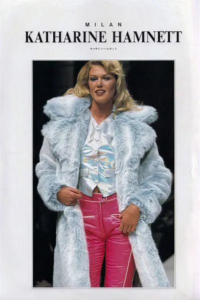 This is a runway or fashion magazine spread for Katharine Hamnett Milan, featuring Japanese text below the designer's name. A tall blonde woman walks the runway wearing a long oversized light blue faux fur coat worn open, a holographic or iridescent multicolored buttoned vest or waistcoat layered over a white button-front shirt, and fitted hot pink vinyl or patent leather trousers with white piping detail along the seams and pockets.