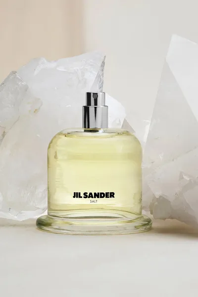 This is a product advertisement photograph showing a glass bottle of Jil Sander Salt perfume with a pale yellow-green liquid inside and a silver spray nozzle, displayed in front of large white crystal or quartz rock formations against a soft beige background.