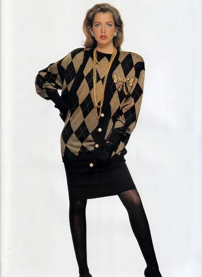 A fashion advertisement or magazine editorial photo showing a woman with blonde hair and blue eyes posing against a plain white background. She is wearing an oversized black and gold argyle-patterned cardigan with large gold buttons, a short black pencil skirt, black opaque tights, black gloves, and black shoes. She is accessorized with layered gold chain necklaces, a gold bow brooch on the cardigan, and gold earrings.