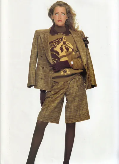 A fashion magazine or advertisement photograph showing a woman with blonde hair wearing a tan and brown plaid double-breasted blazer and matching plaid knee-length shorts, layered over a tan knit sweater featuring a large intarsia horse graphic in yellow and brown. She also wears a brown turtleneck underneath, dark brown tights, burgundy knit gloves, and a watch. The background is plain white.