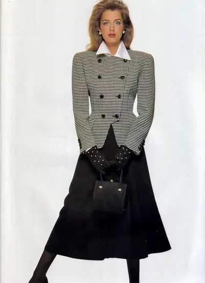 A fashion magazine photograph of a woman with voluminous brown hair and blue eyes, wearing a double-breasted fitted jacket in a black-and-white houndstooth pattern with black buttons, a white collared shirt underneath, a solid black A-line midi skirt, black tights, and black dotted gloves; she is holding a small black structured handbag with gold hardware. The background is plain white.