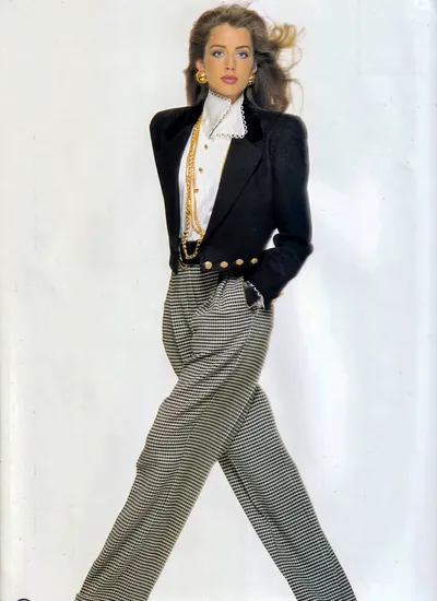 A fashion advertisement or magazine editorial photograph showing a woman with long brown hair and blue eyes, wearing a black blazer with strong padded shoulders over a white ruffled high-neck blouse, layered gold chain necklaces, gold button earrings, a black belt with gold buttons, and black-and-white houndstooth-patterned wide-leg trousers. She is posed against a plain white background with one hand in her pocket.