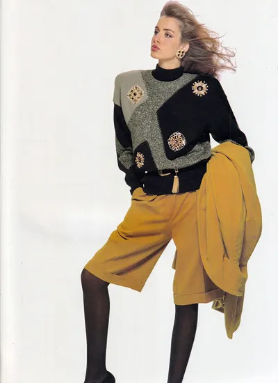 A magazine fashion photograph of a woman with windswept light brown hair, wearing a black and silver metallic colour-block sweater decorated with multiple jewelled brooches, mustard yellow wide-leg bermuda shorts, black tights, and large gold earrings. She carries a matching mustard yellow blazer draped over one arm, and a gold chain tassel belt is visible at her waist.