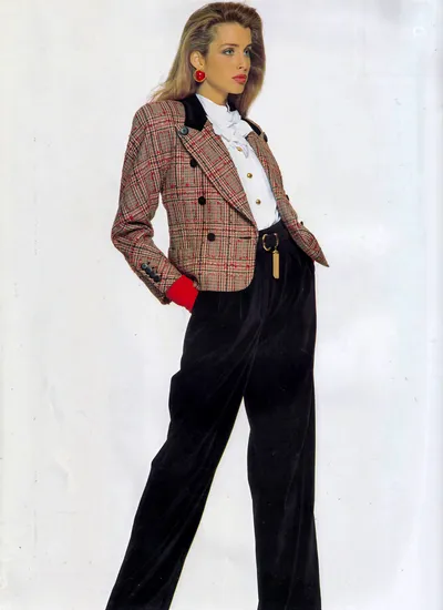 A fashion advertisement or magazine editorial photograph showing a woman with long blonde hair wearing a double-breasted plaid blazer in beige, red, and black with black velvet lapels and red cuffs, paired with a white ruffled blouse and wide-leg black velvet trousers with a gold belt buckle; she also wears large red clip earrings and stands against a plain white background with her hands in her pockets.
