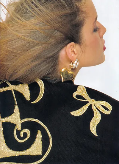A close-up profile shot of a woman with blonde-streaked brown hair pulled back, wearing a large gold heart-shaped drop earring with crystal embellishments at the top. She is wearing a black knit sweater decorated with intricate gold beaded embroidery featuring scrollwork and a bow motif. This appears to be a magazine editorial or advertisement photograph.