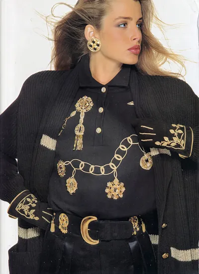 A magazine editorial or advertisement photograph showing a woman with long blonde-brown hair wearing a black ribbed knit open cardigan with gold and silver horizontal stripe accents, layered over a black polo-style knit shirt that is embroidered with gold chain and jewel motifs. She accessorizes with large round gold and black clip-on earrings, black gloves with gold embroidered leaf detailing, a wide black belt with a large gold buckle and attached gold brooch ornaments, and several gold brooches pinned to the shirt.