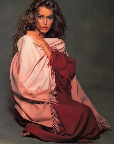 A professional portrait or fashion advertisement photograph of a woman with long wavy brown hair and green eyes, seated on the floor against a muted grey-green background. She is draped in two fringed shawls or wraps — one light blush pink and one deep burgundy-red — layered loosely around her body.