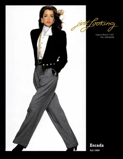 A vintage fashion advertisement from Escada's Fall 1989 collection, sold through a retailer called "Just Looking" in Laguna Beach, California. A woman with long dark brown hair poses against a white background, wearing a black structured blazer with gold button details, a white ruffled high-neck blouse, layered gold chain necklaces, gold earrings, and wide-leg black-and-white houndstooth or small-check patterned trousers.
