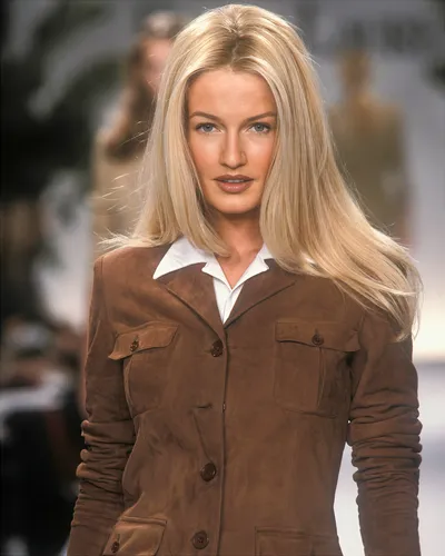 A runway photo of a woman with long blonde hair wearing a brown suede button-front jacket with chest pockets over a white collared shirt; she is walking toward the camera with a blurred runway background behind her.