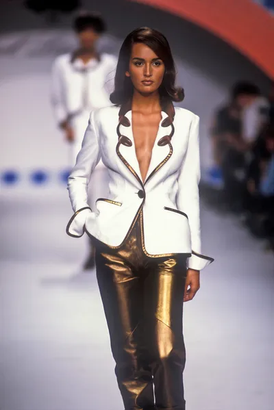 A runway photo showing a woman with dark brown shoulder-length hair walking a fashion show catwalk, wearing a white blazer with dark and gold trim detailing along the deep V-neckline and a single button closure, paired with metallic gold fitted pants. A second model in a white outfit is visible in the blurred background.