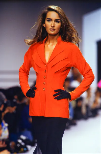 A runway photo of a woman with long brown hair wearing a fitted orange blazer with gold buttons, black gloves, and a dark skirt, walking a fashion show catwalk with a blurred audience in the background.