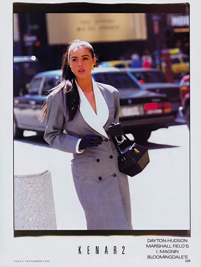 A vintage fashion advertisement from Vogue, September 1990, for the brand Kenar 2, sold at Dayton-Hudson, Marshall Field's, I. Magnin, and Bloomingdale's. A woman with long dark hair pulled back is photographed outdoors on a city street. She wears a grey houndstooth double-breasted longline jacket with a wide white lapel or scarf, paired with what appears to be a matching grey houndstooth skirt. She carries a structured black handbag with gold hardware and wears black gloves and yellow gold earrings. Cars and urban buildings are visible in the background.