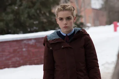A young woman with blonde braided hair wearing a dark red coat over a school uniform with a light blue collar and dark tie, standing outdoors in a snowy setting near a brick wall. This appears to be a production still or promotional photo from a film or television series.