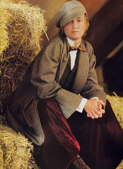 This appears to be a fashion advertisement or editorial photograph featuring a young woman with reddish-blonde hair sitting against hay bales in what looks like a barn setting. She is wearing a grey herringbone flat cap, a grey herringbone oversized jacket with wide lapels, a white shirt with a floral or patterned necktie, a grey herringbone waistcoat, and dark red or burgundy velvet trousers, with lace-up ankle boots partially visible at the bottom.