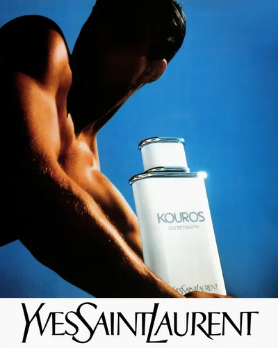 This is a fragrance advertisement for Yves Saint Laurent's Kouros Eau de Toilette. It shows a shirtless, muscular man with dark hair leaning forward and holding a white rectangular perfume bottle with silver metallic clip details against a bright blue background. The brand name "Yves Saint Laurent" appears in large stylized text along the bottom of the image.