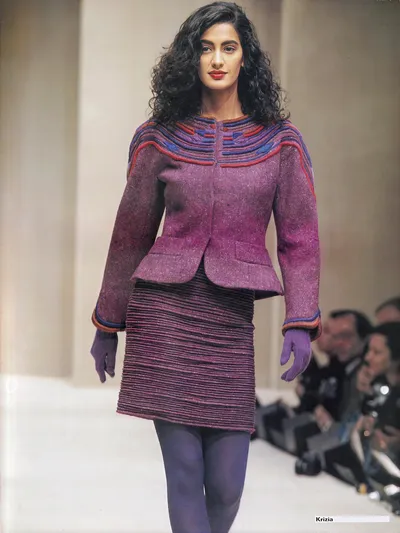 A runway photo showing a woman with long dark curly hair and red lipstick walking on a white catwalk. She wears a fitted purple-mauve tweed jacket with decorative ribbed trim in purple, red, and blue along the neckline and cuffs; a separate dark ribbed pencil skirt in brown-purple horizontal stripes; purple gloves; and purple tights. The brand name "Krizia" is printed in the bottom right corner.