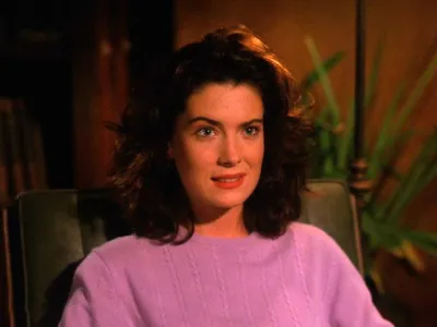 A still from what appears to be a film or TV production, showing a young woman with shoulder-length dark curly hair and red lipstick, wearing a lavender cable-knit sweater, seated in a chair and looking toward the camera with a slight smile. A green plant is visible in the warm-toned background.