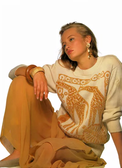 A fashion or editorial photograph of a young woman with light brown hair pulled back, seated on the floor against a plain white background. She is wearing a cream knit sweater with a large golden-orange graphic print featuring what appears to be an abstract or Aztec-style bird or figure, paired with loose mustard-yellow wide-leg trousers in a sheer or lightweight fabric. Her accessories include large ornate drop earrings, a chunky brown wooden bangle, and a gold cuff bracelet worn on the same wrist. She is barefoot and is looking upward and to one side.